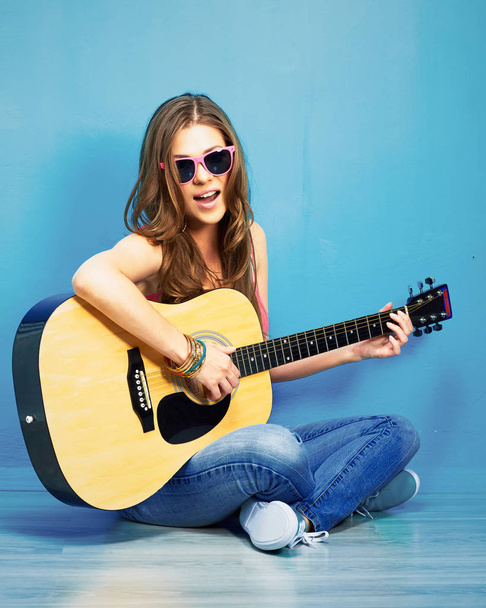 fashion music look with young model in sunglasses sitting on blue floor and playing acoustic guitar  - Zdjęcie, obraz