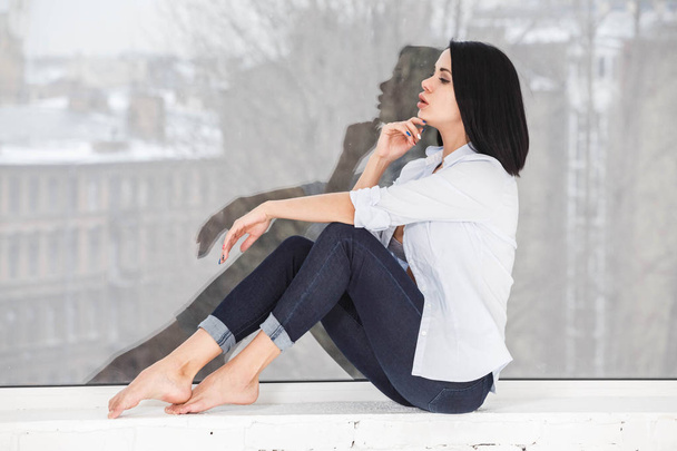 Studio shot of beautiful brunette woman wearing dark jeans and white shirt sitting on windowsill near window at apartment interior - Foto, imagen