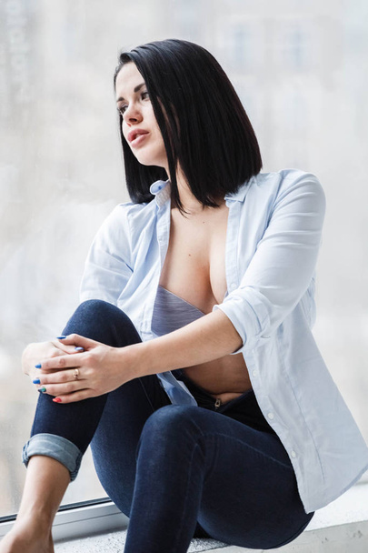 Studio shot of beautiful brunette woman wearing dark jeans and white shirt sitting on windowsill near window at apartment interior - Фото, изображение
