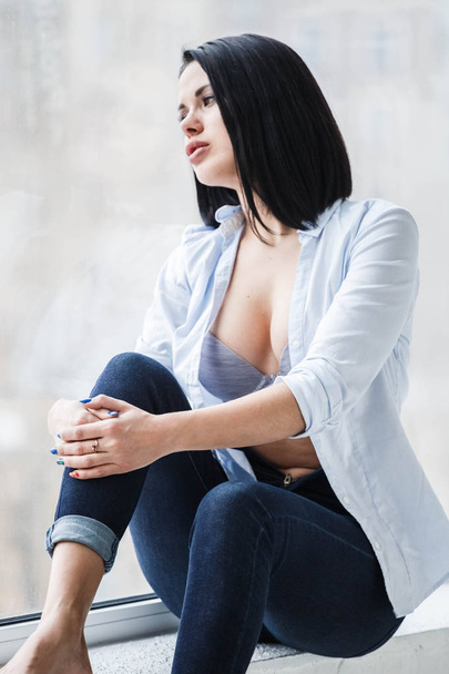 Studio shot of beautiful brunette woman wearing dark jeans and white shirt sitting on windowsill near window at apartment interior - Foto, Bild