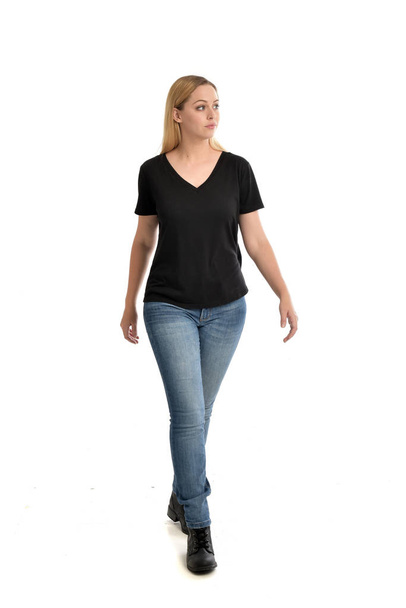 Ganzkörperporträt eines blonden Mädchens, das ein einfaches schwarzes Hemd und Jeans trägt und stehend auf weißem Hintergrund posiert. - Foto, Bild