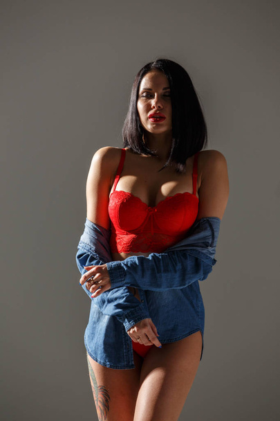Studio záběr krásná bruneta žena nosí sexy červené spodní prádlo. Mladá kráska žena s tetování na tělo stojící v parkovacím světlem na pozadí šedé studio  - Fotografie, Obrázek