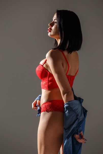 Studio shot of beautiful brunette woman wearing sexy red lingerie. Young beauty woman standing in side light on grey studio background  - Photo, image