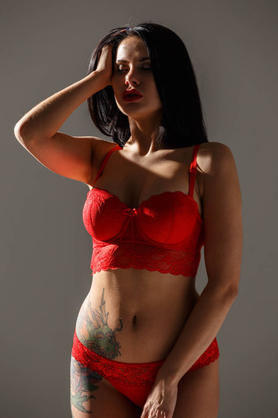 Studio shot of beautiful brunette woman wearing sexy red lingerie. Young beauty woman with tattoo on body standing in side light on grey studio background  - 写真・画像