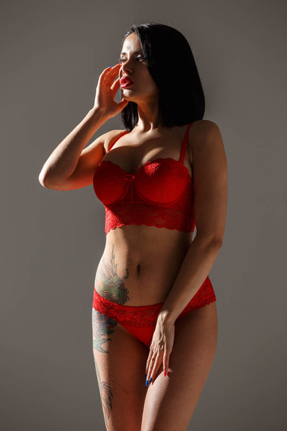 Studio shot of beautiful brunette woman wearing sexy red lingerie. Young beauty woman with tattoo on body standing in side light on grey studio background  - Photo, Image
