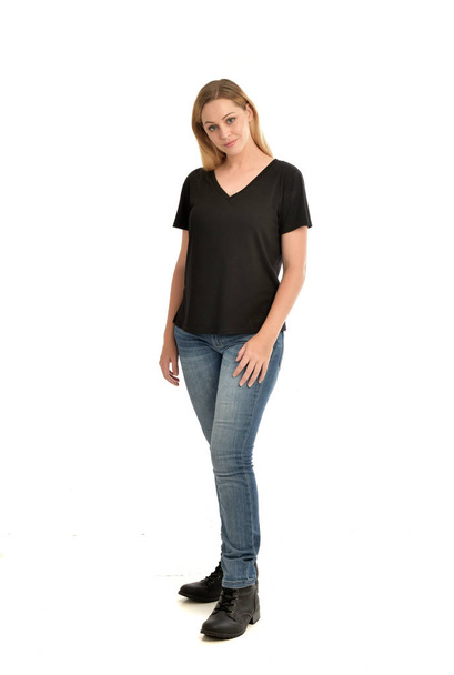 Ganzkörperporträt eines blonden Mädchens, das ein einfaches schwarzes Hemd und Jeans trägt und stehend auf weißem Hintergrund posiert. - Foto, Bild