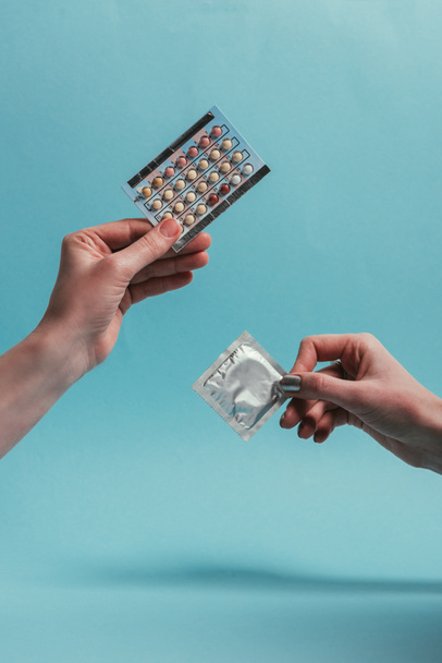 partial view of women holding condom and contraceptive pills in hands isolated on blue - Fotoğraf, Görsel