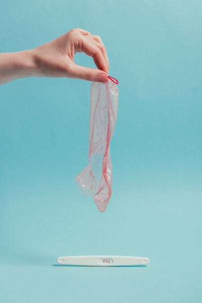 cropped shot of woman holding ripped condom in hand and pregnancy test isolated on blue - Photo, Image