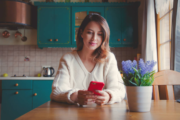 πορτρέτο του Κοκκινομάλλης γυναίκα με κινητό τηλέφωνο που κάθεται στο τραπέζι στην κουζίνα.  - Φωτογραφία, εικόνα