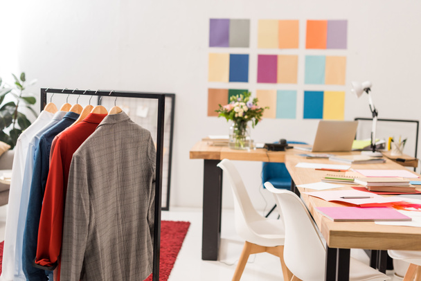 μοντέρνα ρούχα σε κρεμάστρες, χαρτιά στο τραπέζι στο σύγχρονο γραφείο με χρωματική παλέτα στον τοίχο - Φωτογραφία, εικόνα