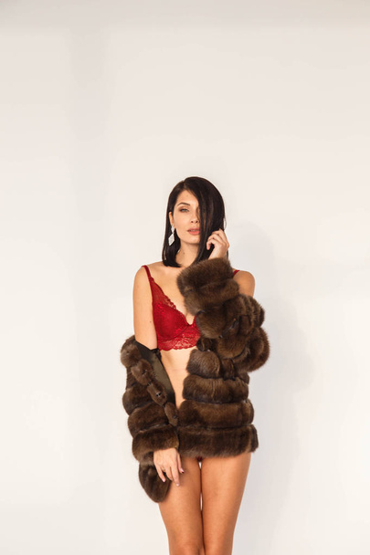 Studio portrait of beautiful brunette girl in sexy red lingerie. Shot of young woman in fur coat standing on white background - Φωτογραφία, εικόνα