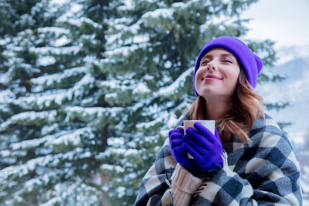 portrait of redhead girl in plaid and purple hat with cup of hot drink at winter outdoor - Photo, Image