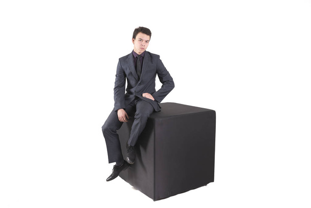 Un joven con un traje de negocios sentado en un cubo negro aislado sobre fondo blanco
 - Foto, Imagen