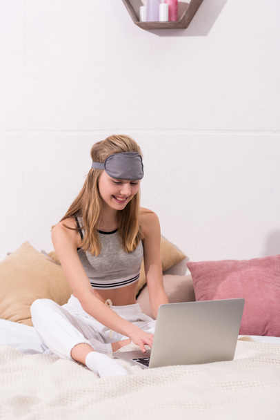 kaunis nuori nainen, jolla on unimaski päähän työskentelemään kannettavan tietokoneen kanssa, kun hän istuu sängyssä aamulla
 - Valokuva, kuva