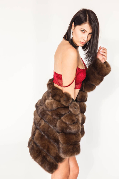 Studio portrait of beautiful brunette girl in sexy red lingerie. Shot of young woman in fur coat standing on white background - Foto, Bild