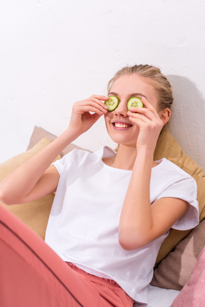 happy young woman applying cucumber slices on eyes at home - Fotoğraf, Görsel