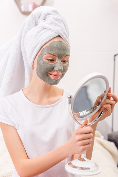 jeune femme avec masque en argile cosmétique regardant miroir à la maison
 - Photo, image