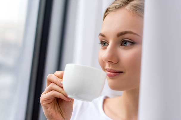 smiling young woman drinking coffee and looking through window - Foto, Imagem