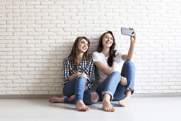 Portrait of two millennial females, fooling around in front of mobile smartphone camera. Brown eyed model girls with long brunette wavy hair and beautiful smiles in white blank and checkered shirt. - Foto, Imagem