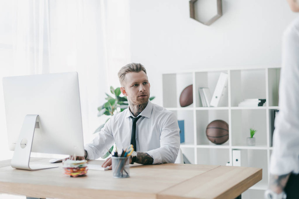 handsome young tattooed businessman looking away while working with desktop computer in office - Photo, Image