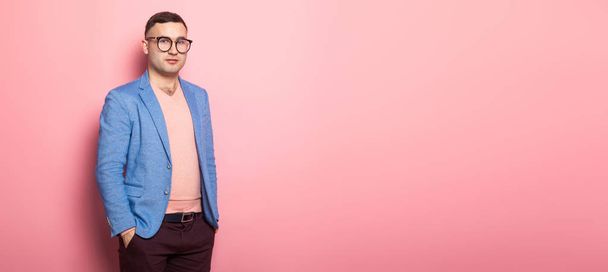 Handsome young man in blue jacket holding hands in pockets on pink background  - Φωτογραφία, εικόνα