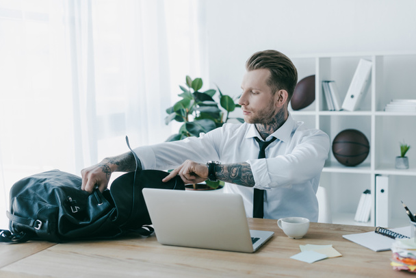 young tattooed businessman opening backpack while working with laptop at workplace - Foto, Bild