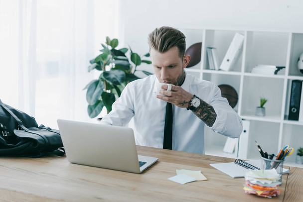 handsome young businessman drinking coffee and using laptop at workplace - Foto, Bild