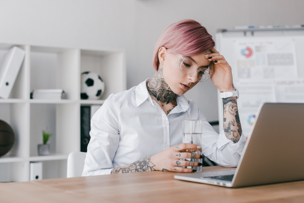 cansada joven empresaria sosteniendo un vaso de agua y usando un portátil en la oficina
 - Foto, Imagen