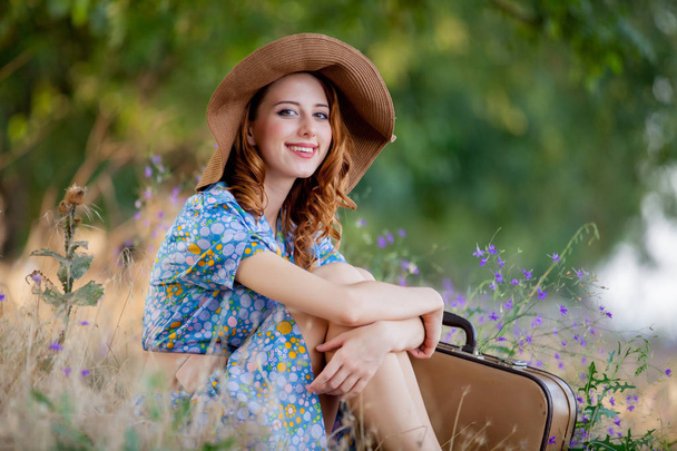 retrato de mujer joven en vestido azul en sombrero con maleta sentado en la hierba en el campo al aire libre
 - Foto, imagen