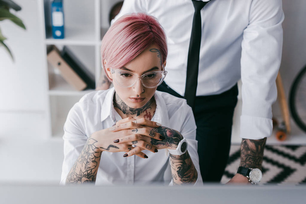 fokussierte junge Geschäftsfrau sitzt am Arbeitsplatz, während männlicher Kollege hinter ihr steht - Foto, Bild
