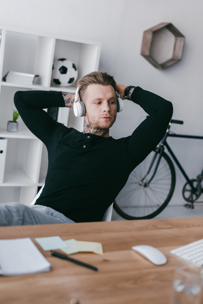 guapo joven tatuado hombre en auriculares sentado con las manos detrás de la cabeza en el lugar de trabajo
  - Foto, imagen