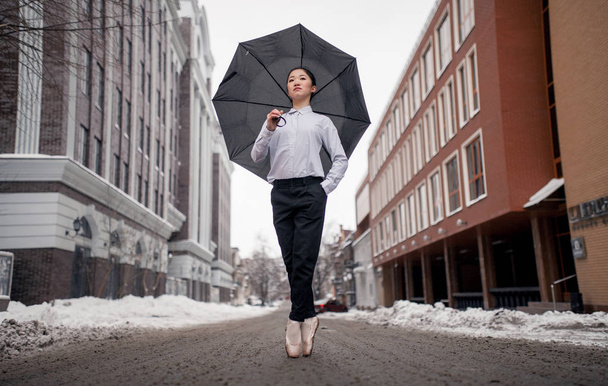 Ballerina with umbrella in her hands is standing at city street against snow and buildings background. - Photo, Image