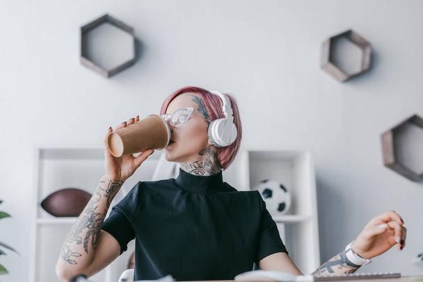 young businesswoman in headphones drinking coffee from paper cup at workplace - Фото, зображення