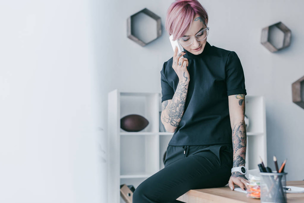 glimlachend jonge zakenvrouw met tatoeages praten door de smartphone op werkplek - Foto, afbeelding