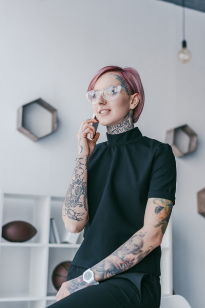 smiling young tattooed businesswoman talking by smartphone and looking away at workplace - Фото, изображение