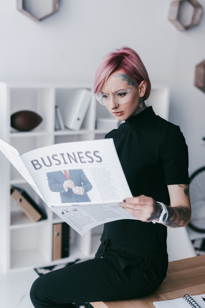 serious young tattooed businesswoman in eyeglasses reading newspaper at workplace - Photo, Image