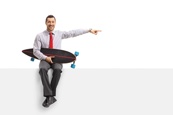 Businessman with a longboard sitting on a panel and pointing isolated on white background - Fotografie, Obrázek