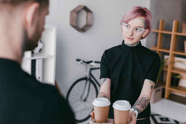 selective focus of young tattooed woman giving paper cups to colleague - Foto, Imagen