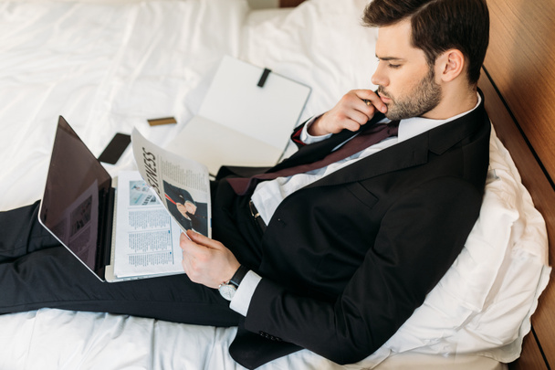 high angle view of businessman lying on bed in hotel room and reading newspaper - Photo, Image