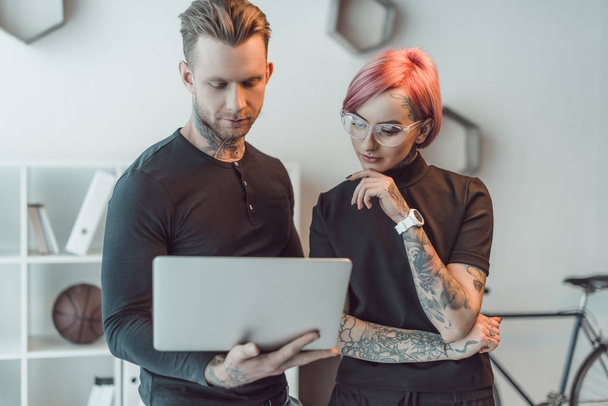young tattooed colleagues using laptop together in office - Фото, зображення