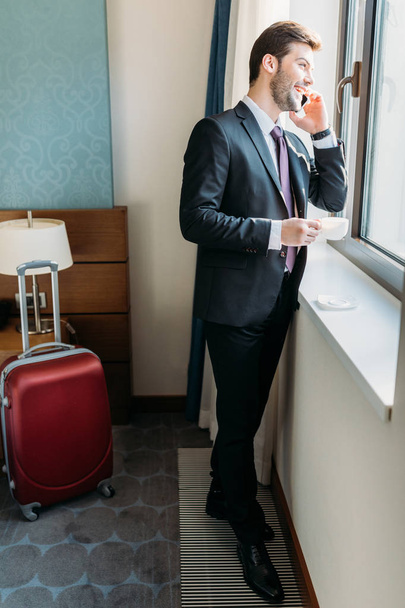 knappe zakenman praten door smartphone in hotelkamer en kijken naar venster - Foto, afbeelding