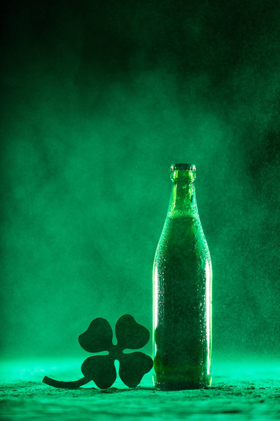 Wet green beer bottle and a shamrock standing on a dusty background. St. Patrick's Day. - Photo, Image