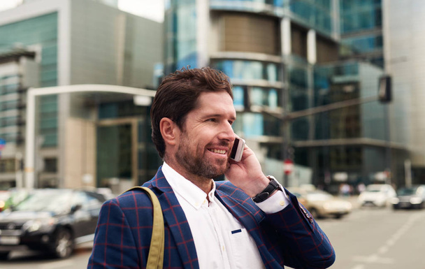 Lächelnder junger Geschäftsmann im Blazer, der auf der Straße in der Stadt unterwegs ist und sich mit dem Handy unterhält   - Foto, Bild