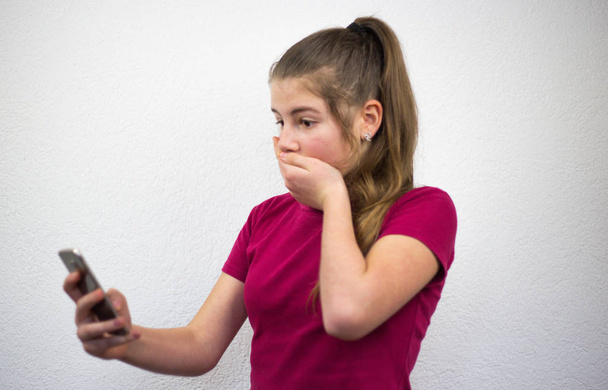Joven linda adolescente rubia sorprendida por su teléfono inteligente móvil mostrando emoción y cubriéndose la boca con la mano usando camiseta rosa sobre un fondo blanco
 - Foto, Imagen