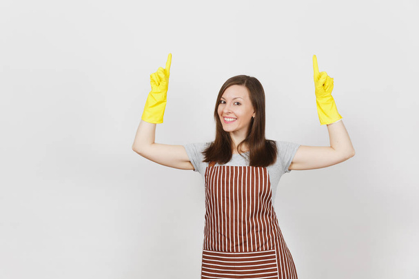 Young attractive smiling housewife in striped apron, yellow gloves isolated on white background. Beautiful housekeeper woman pointing index fingers up looking camera. Copy space for advertisement. - Photo, Image