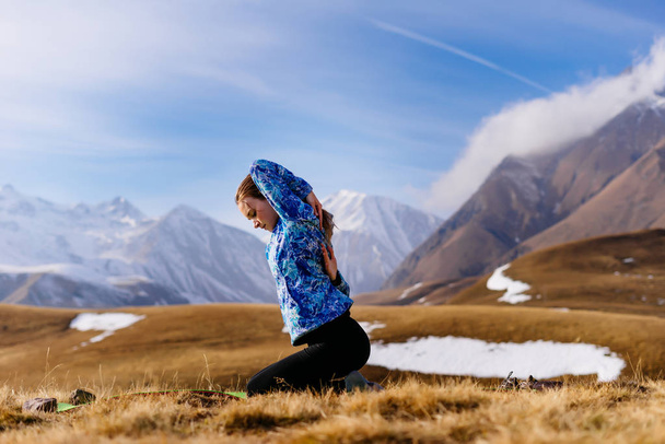 active young girl in a blue jacket doing yoga in the background of the Caucasian mountains - Photo, Image