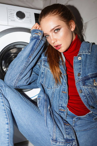 Retrato de mujer joven en chaqueta de mezclilla mirando cuidadosamente en cámara con lavadora en el fondo
 - Foto, Imagen