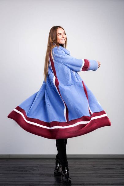 Young dark-haired woman in blue fur coat made from natural mink fur with claret-colored white stripes is relaxed, posing and dancing on white isolated background - Photo, Image