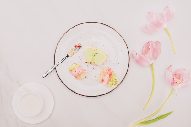 top view of pieces of cake on plate with fork, flowers and cup of milk on tabletop  - Foto, afbeelding