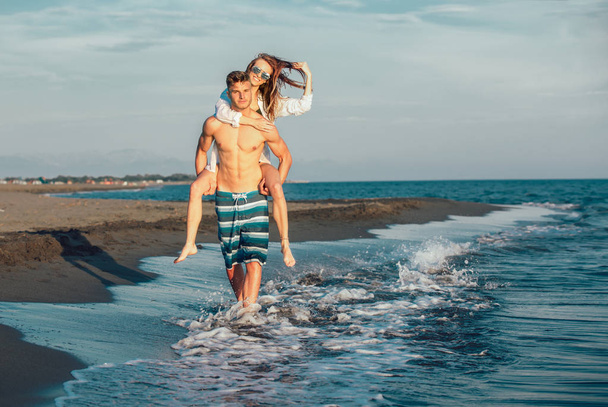 Ευτυχισμένο ζευγάρι στην αγάπη στην παραλία καλοκαιρινές διακοπές. Χαρούμενο κορίτσι piggybacking σχετικά νεαρό φίλο διασκεδάζοντας. - Φωτογραφία, εικόνα